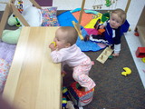 Mary standing at the bench in daycare with her feet inside a cloth box trying to lift her left foot out.