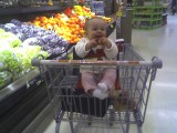 Mary sitting in a shopping cart with her legs straight out and a big smile.  You can see fruit in the background.