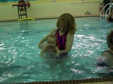 Mommy is holding Mary under her arms in the pool as if she is swimming and Mary is splashing both hands in front of her.