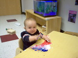 Mary doing some finger painting.  She is in a blue jersey and has a big smile on her face as she makes a mess on the table.