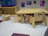 Mary under a little kids table on her belly.  She is peering out and you can only see her face.
