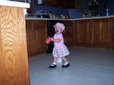 Mary in the same pink little school girl outfit and a pink waif hat. She is walking by a gallon container of milk with a sippy cup of milk in her hand.