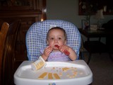Mary sitting in her highchair with yogurt all over her face.  She has her spoon in her mouth like a cigar.