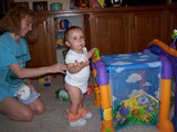 Mary standing at her toy, her upper body turned towards the camera and her mouth open talking to it.  Her pants are down and her onesie is unsnapped and her diaper is off.  Mommy is behind her.